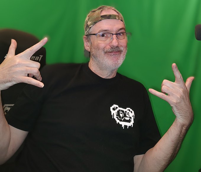 A bearded man wearing a camouflage cap backwards, glasses, and a black t-shirt makes hand gestures against a green screen background, with a microphone partially visible on the left.