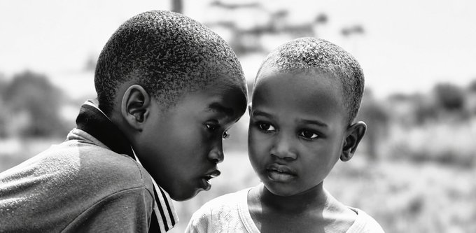 A black-and-white portrait of two young African children, a boy in profile and a girl looking forward with a subtle expression, set against a blurred outdoor background.