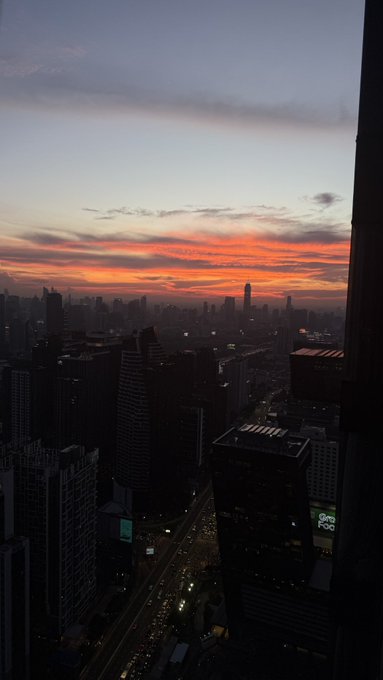 A high-rise view of a city skyline at sunset, featuring silhouetted skyscrapers against a vibrant orange and pink sky.
