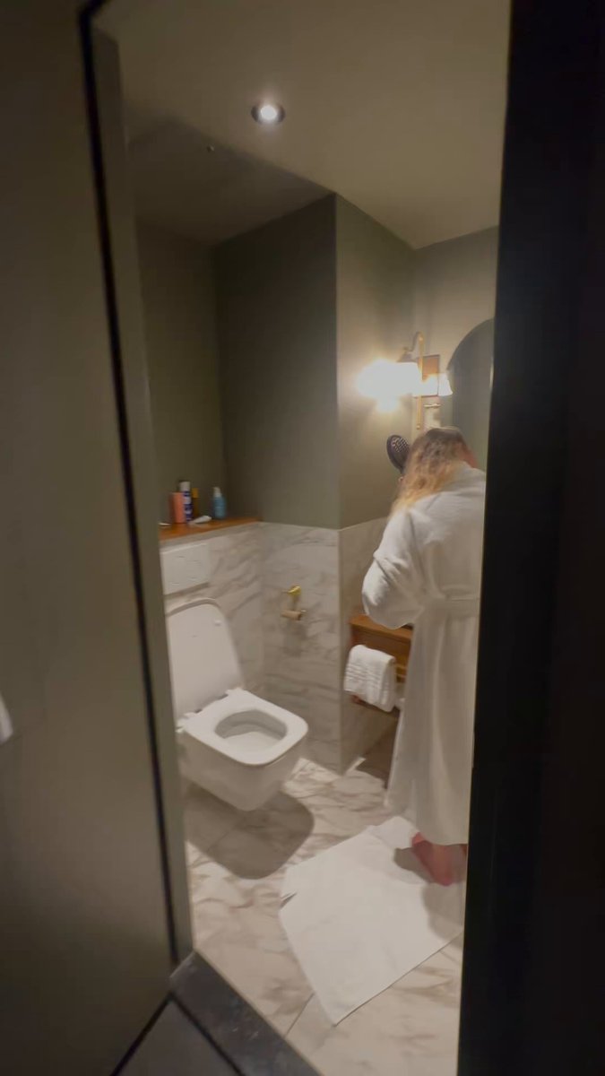 The image shows a person standing in front of a bathroom mirror, wearing a white robe or bathrobe. The bathroom features a toilet with a closed lid and a sink with a towel hanging from it. There are also several bottles placed on the countertop near the sink. The overall scene suggests that this is likely a hotel room, as indicated by the post about hotels losing money on perpetuals.