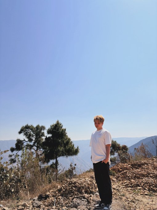 A young man with light hair wearing a white t-shirt and black pants standing on a rocky overlook with mountains and a clear blue sky in the background.