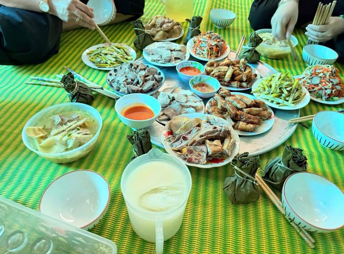 A top-down view of a traditional Vietnamese feast laid out on a yellow and green striped mat, featuring numerous dishes including chicken, vegetables, dipping sauces, and what appears to be banh tet (sticky rice cakes wrapped in leaves).