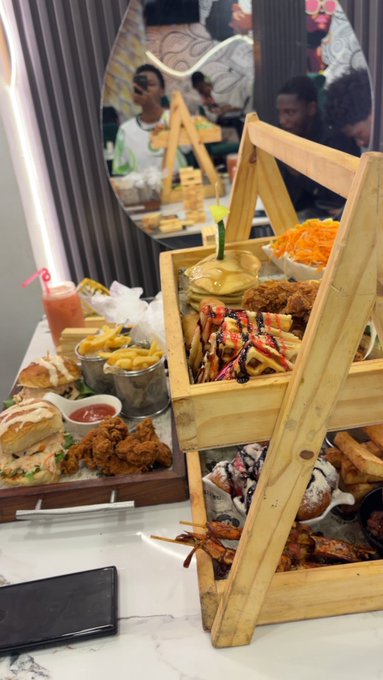 A wooden tiered serving stand loaded with a feast of fried chicken, fries, sandwiches, and desserts displayed on a marble countertop at a restaurant.