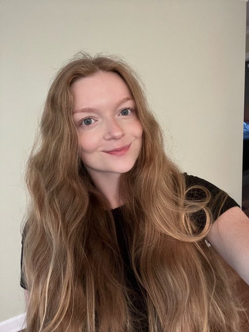 A selfie of a young woman with long reddish-brown hair, fair skin, and freckles, smiling at the camera against a plain wall.