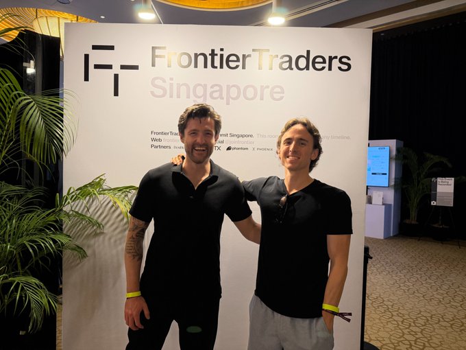 A photograph of two smiling men in black t-shirts standing in front of a 'Frontier Traders Singapore' event backdrop, with the Frontier Traders logo and partner logos visible.