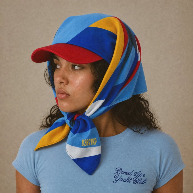 A woman models a colorful red, blue, yellow, and white striped cap and matching scarf, with "BZ2021NA" visible on the scarf and "Bored Ape Yacht Club" text on her light blue t-shirt, tying into the post's promotion of Bored Ape Yacht Club's "Manifest 001: Flying Colors" collection and upcoming Meet at the Clubhouse event.