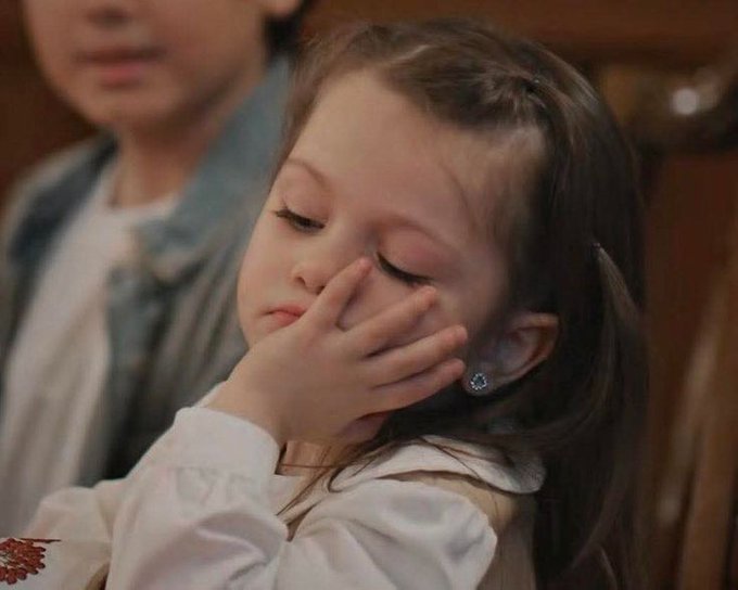 A young girl with braided hair resting her face against her hand with eyes closed, appearing tired or contemplative, with a person in a denim jacket visible in the background.