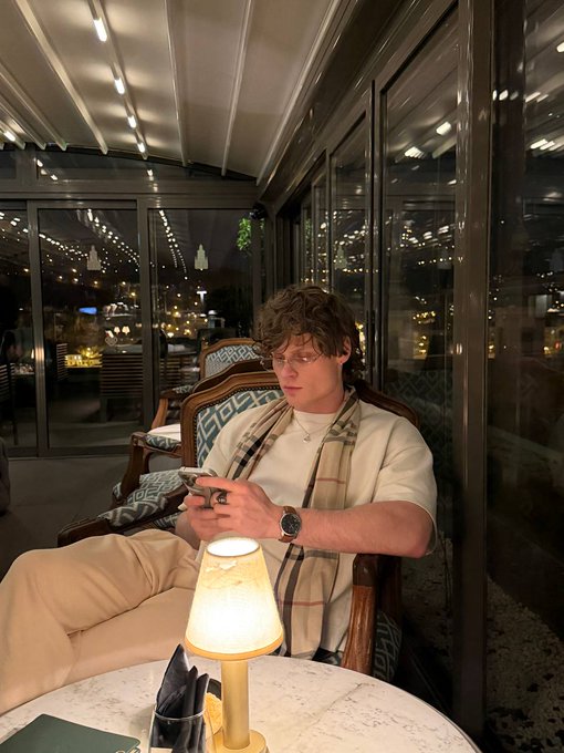A young man with glasses and a Burberry scarf sits on a chair in a stylish cafe or lounge, looking at his phone with a small table lamp in the foreground.