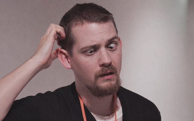 A man with a goatee scratching his head and looking to the side with a puzzled or confused expression.