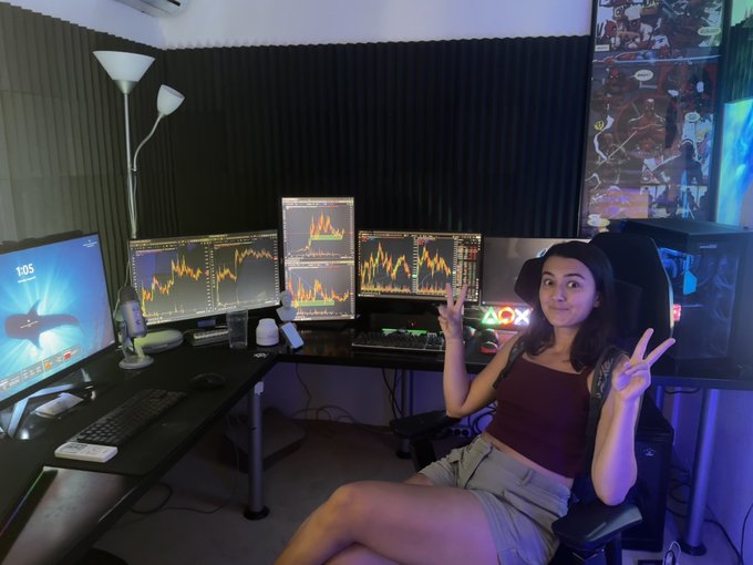 Young woman sitting in a gaming/trading chair in a room with multiple monitors displaying financial charts, making peace signs with both hands, with comic book artwork on the walls and RGB lighting.