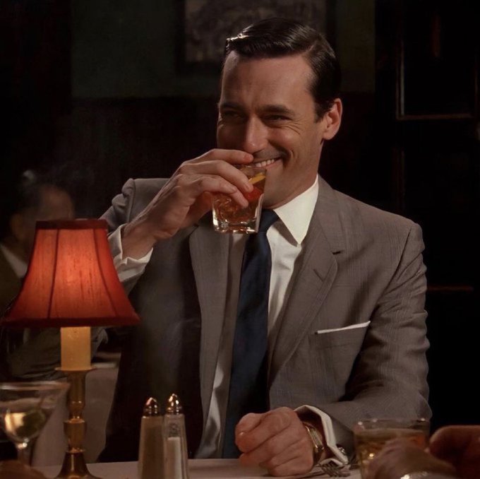 Don Draper from the TV series Mad Men, wearing a grey suit and blue tie, smiles while holding a glass of whiskey in a dimly lit lounge setting.