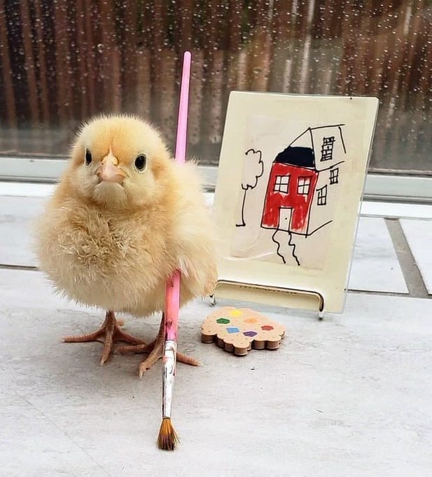 A small, fluffy yellow chick stands holding a large pink paintbrush in its wing, positioned next to a small canvas displaying a child-like painting of a red house, with a flower-shaped paint palette nearby.