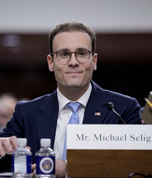 A man in a navy suit and glasses, identified by a nameplate as 'Mr. Michael Selig', sits at what appears to be a congressional or formal hearing table with water bottles in front of him.