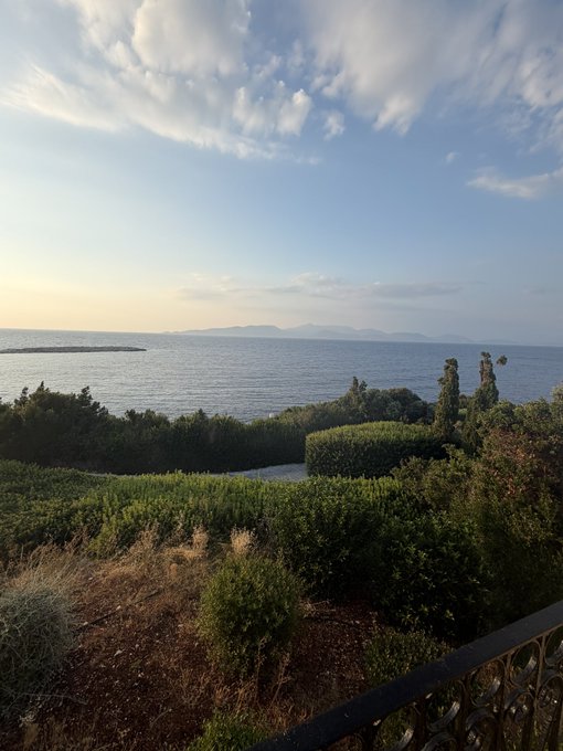 A serene coastal landscape with lush greenery in the foreground opens up to a calm sea and distant mountains under a soft, partly cloudy sky, capturing the peaceful beauty of a new day. Fits perfectly with a simple "Gm." greeting to share a tranquil morning view.