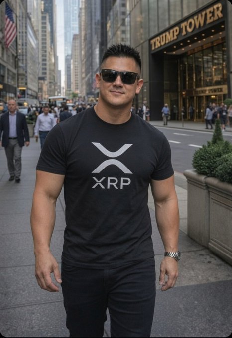 A muscular man wearing sunglasses and a black "XRP" t-shirt poses confidently on a Manhattan sidewalk in front of Trump Tower, tying into his playful announcement about running for California Senate while sporting the popular crypto symbol.