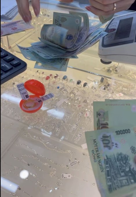 Hands count Indonesian Rupiah banknotes (100,000 denomination) at a glass display counter showcasing rings and jewelry, with a red ring box visible.