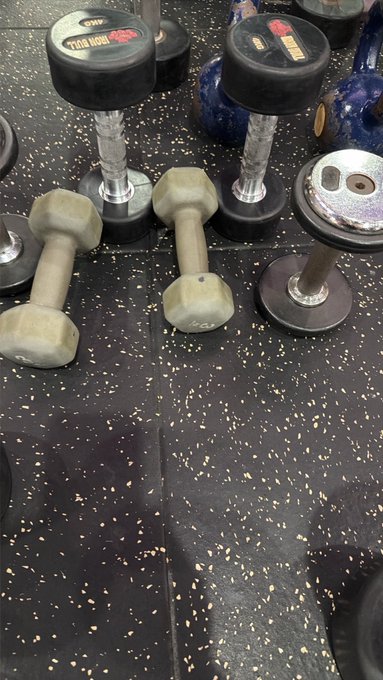 Several dumbbells of various sizes scattered on a black rubber gym floor with white speckles.