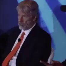 The image features a man wearing an orange tie, sitting down with his hands together. He appears to be engaged in conversation or observing something happening around him. The background of the image shows a blue wall, which contrasts with the man's attire and adds depth to the scene. The caption "Watching certain long-time $MSTR bulls begin to doubt:". This phrase is likely an inside joke or reference to a recent event involving bulls in the context of the conversation between the man and someone else present in the image.
