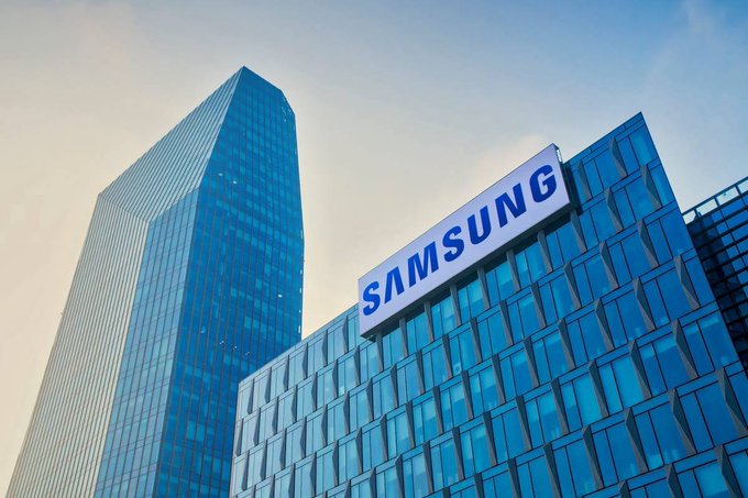 A low-angle view of a tall glass skyscraper next to a building displaying a large 'SAMSUNG' sign against a blue sky.