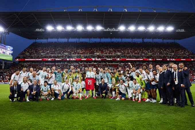 A celebratory team photo of a football squad and coaching staff posing on the pitch with a '21' jersey displayed, taken in a stadium at night with fans in the stands.