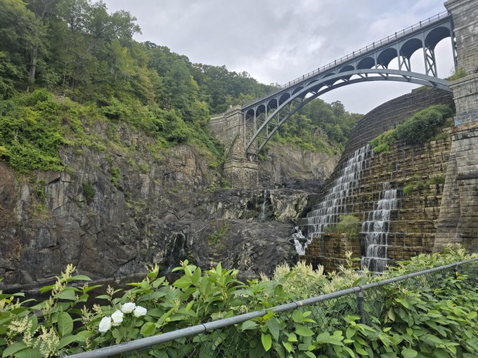 A scenic view of a stone arch bridge spanning a gorge, with a cascading waterfall flowing down the rocky cliffside and lush greenery in the foreground.