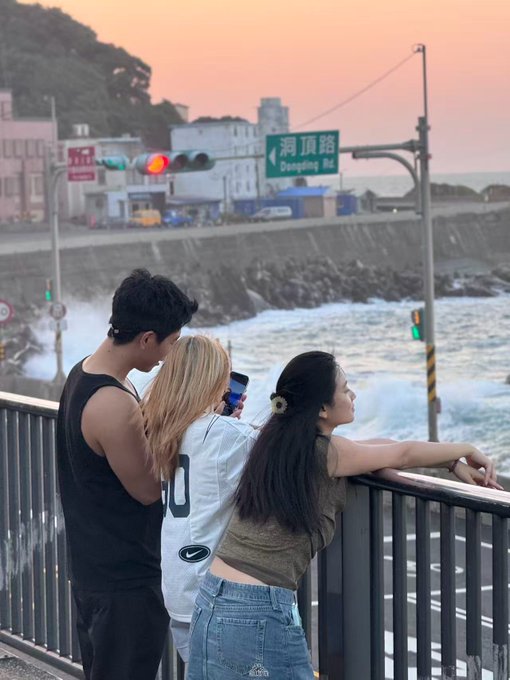 Three young people lean against a railing along a coastal road at sunset, with a green street sign reading '洞頂路 Dongding Rd' visible overhead.