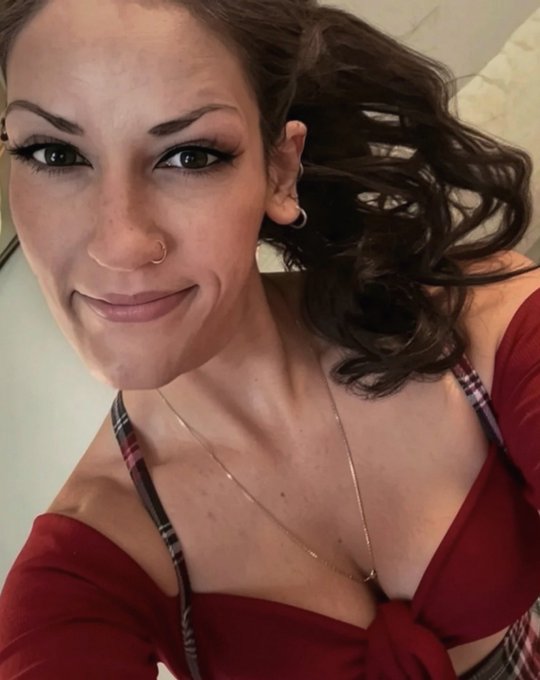 A selfie of a smiling woman with dark hair, a nose ring, and wearing a red top with plaid straps.