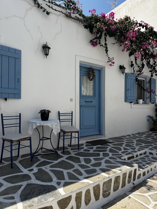A white-washed building with blue doors, blue shutters, and a stone-paved patio is decorated with cascading bougainvillea flowers and a small table with two blue chairs.