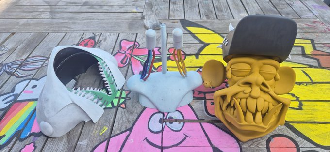 Three sculptural objects rest on a graffiti-covered wooden surface: a white shark-like helmet with teeth, a small central piece with wires and candles, and a tan monkey-like face wearing a black cap.