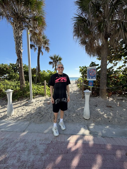 A person standing on a beach path between palm trees wearing sunglasses, a black TRILLSTAR t-shirt, black shorts, and white sneakers, with the ocean visible in the background.