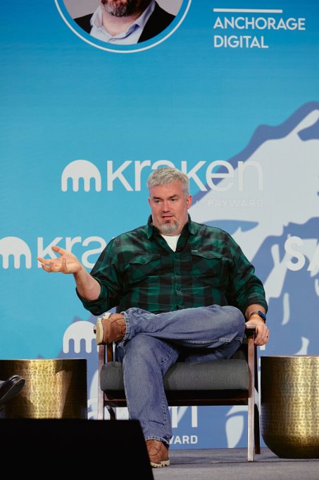 A gray-bearded man in a green and black plaid shirt speaks animatedly while seated on stage at the SALT Conference, with "ANCHORAGE DIGITAL," "Kraken," and "JOBY" branding visible on the backdrop behind him.