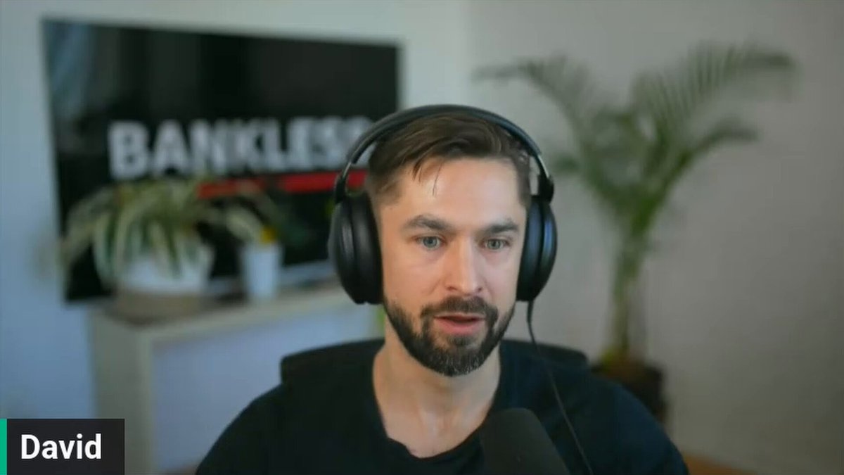 The image features a man wearing headphones while sitting at a desk with a computer monitor behind him. The man appears to be talking into a microphone or headset, possibly participating in an online meeting or recording a video call. The background of the image includes a TV screen and a potted plant, suggesting that he might be working from home or in a casual office environment.