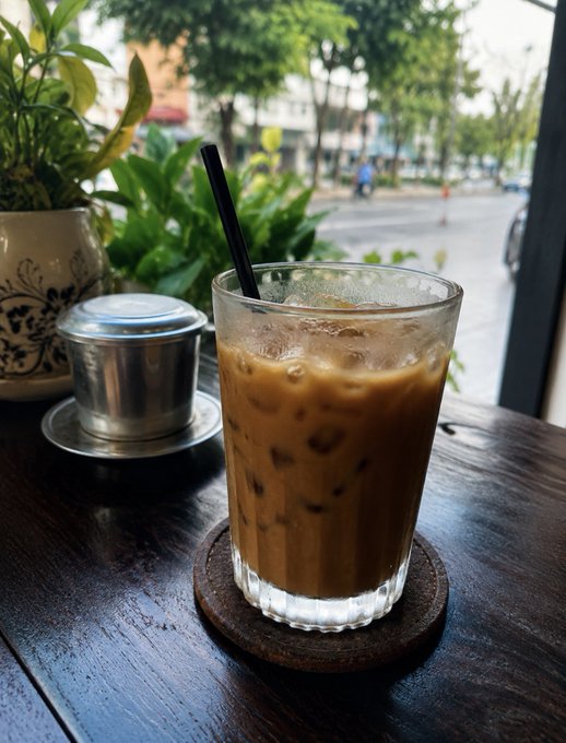A glass of Vietnamese iced coffee with a black straw sits on a wooden table next to a traditional Vietnamese coffee filter (phin), with potted plants and a street view in the background.
