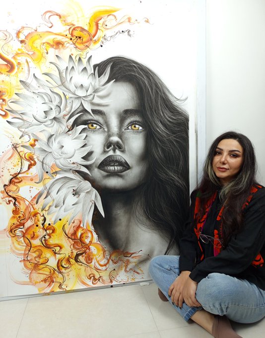 A woman with dark hair sits cross-legged on the floor in front of a large portrait painting featuring a woman's face surrounded by white flowers and orange watercolor swirls.