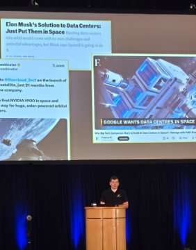 A presenter stands at a podium in front of a large screen displaying articles titled 'Elon Musk's Solution to Data Centres: Just Put Them in Space' and 'Nvidia wants Data Centres in Space'.