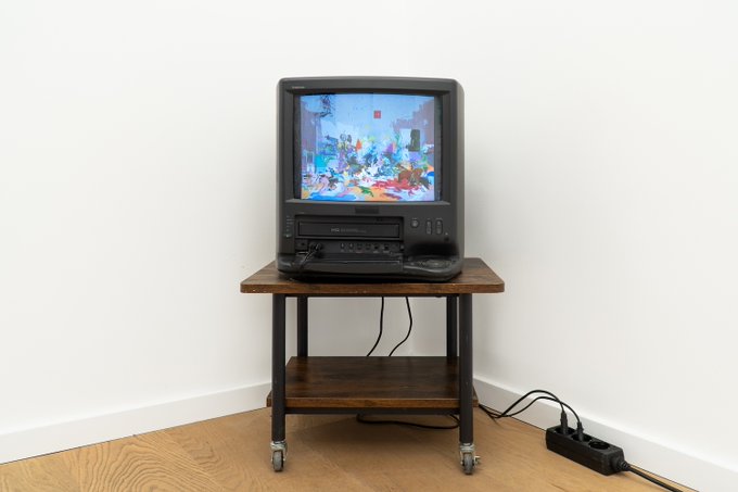 A vintage CRT television and VCR setup on a wooden rolling cart displays a colorful digital artwork featuring abstract forms and characters, with cables running to a power strip on the floor—likely presenting the NFT pieces available on objkt.com through a retro-tech medium.