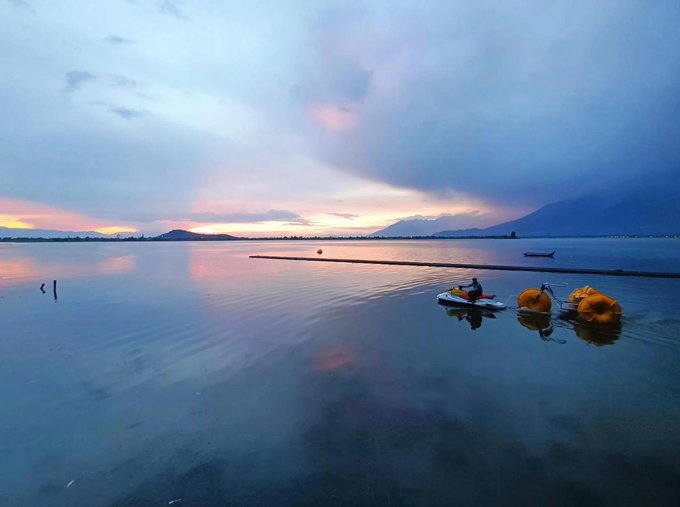 A lone figure on a paddleboard sits on a mirror-still lake at sunset, with calm water reflecting the pink and orange sky, creating a serene moment of quiet presence amid vast open space—mirroring the post's message of confidently taking up space and owning one's presence in the world.