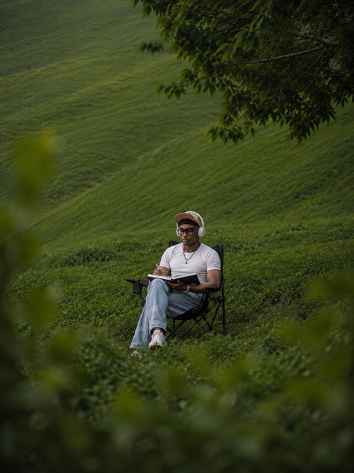 A man wearing a cap, white t-shirt, jeans, and white headphones sits in a black folding camping chair on a lush green hillside, reading a book with trees framing the scene.