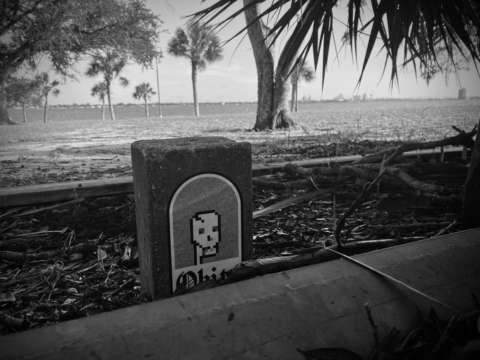 A black-and-white photograph of a stone grave marker with a pixel art skull and the partially visible word "Ohit", set in a coastal landscape with palm trees.