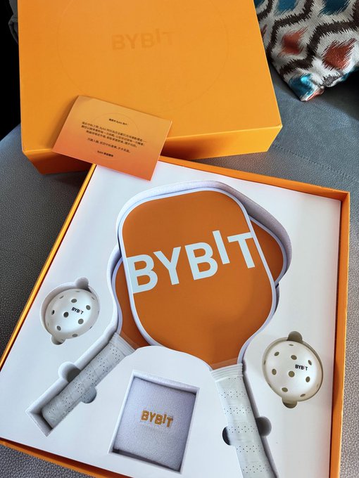 A Bybit-branded orange pickleball set, including two paddles, two balls, and a carrying case, displayed in its matching orange gift box. The accompanying post expresses gratitude to Bybit_ZH and JohnnyBybit for the Mid-Autumn Festival gift, playfully challenging other crypto exchanges to follow suit.