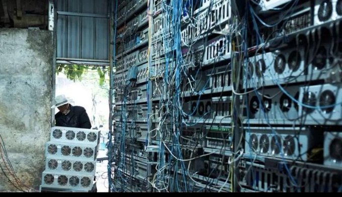 A person wearing a hat stands amidst a massive, tangled installation of electronic equipment and hanging cables.