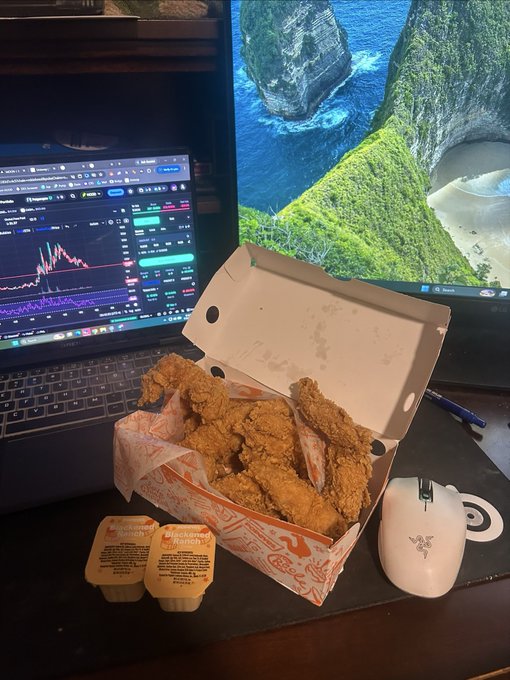 A box of Popeyes chicken tenders sits on a desk in front of a laptop showing trading charts and a monitor displaying a tropical landscape, with two sauce containers labeled "BoldBQ" and "Blackened Ranch" beside it—paired with the post's $TENDIES reference, humorously framing the fried chicken as both the meal and the meme stock ticker.