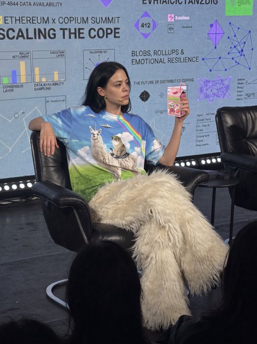 A woman sits on stage at the "Ethereum x Copium Summit" panel titled "Scaling the Cope: Blobs, Rollups & Emotional Resilience," wearing a llama-print shirt and draped in a large furry llama costume while holding a Japanese drink carton.

The image fits the "could've been an EIP" joke perfectly—the "Copium Summit" backdrop, with its tongue-in-cheek Ethereum jargon ("EIP-4844 Data Availability," "Rollup Settlement," "Execution Layer"), parodies serious technical Ethereum Improvement Proposals, and the absurd llama mascot visually embodies the copium theme of coping with crypto disappointments rather than proposing real upgrades.