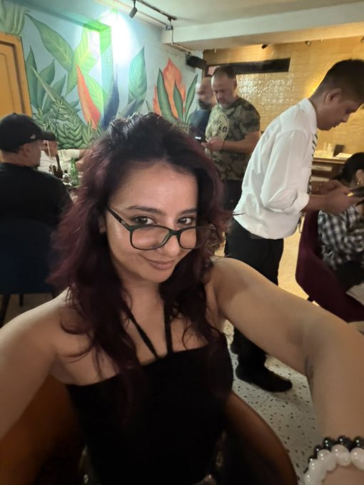 A woman with dark reddish hair and glasses takes a selfie in what appears to be a restaurant, while a waiter in a white shirt and other patrons are visible in the background.