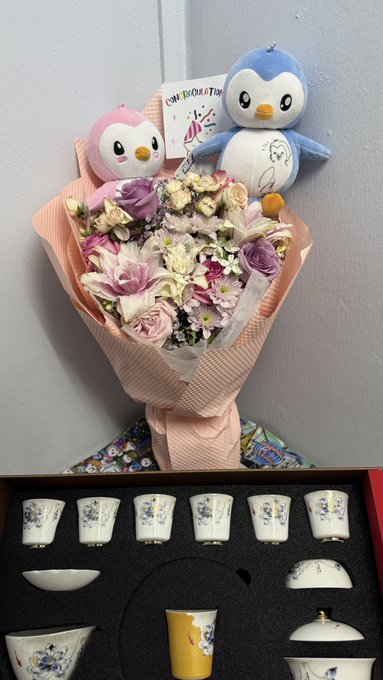 A bouquet of pastel flowers wrapped in pink paper, decorated with two penguin plush toys and a 'Congratulations' card, displayed above a boxed tea set with floral designs.