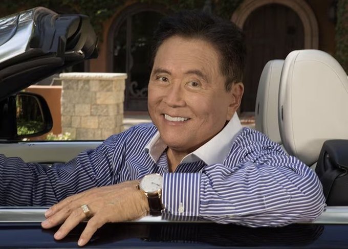 An older smiling man in a striped blue shirt leaning out of a convertible car, wearing a watch and a ring, with a home visible in the background.