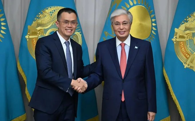 Two men in business suits shaking hands in front of Kazakhstan flags, appearing to be officials at a formal diplomatic meeting.
