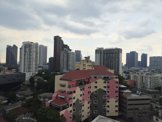 An elevated cityscape view showing modern high-rise buildings in the background and a distinctive pink, black, and yellow camouflaged building with a red roof in the foreground under a cloudy sky.