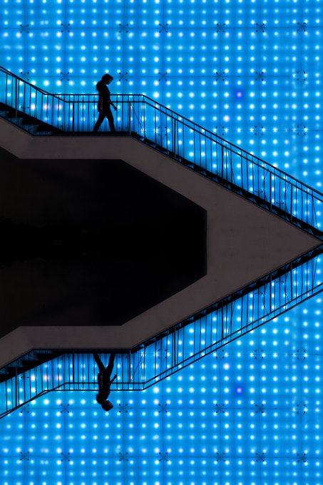 A silhouette of a person walks down a modern zigzagging staircase against a backdrop of bright blue illuminated panels arranged in a grid pattern, creating a striking geometric composition.