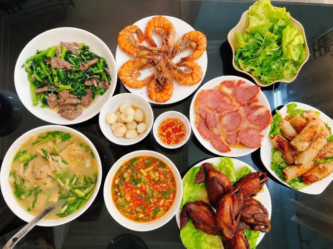 A Vietnamese family-style dinner spread featuring dishes like stir-fried beef with greens, fried shrimp, sliced ham, fried spring rolls, roasted pigeon, fresh herbs and lettuce, quail eggs, chili dipping sauce, and a bowl of soup with chicken.

The image pairs with the post's warning to "enjoy dinner but stay cautious" — a reminder that just as a full meal can be satisfying, overextending after a strong market rally can lead to a sharp pullback, so investors should eat, then think clearly before taking profits.
